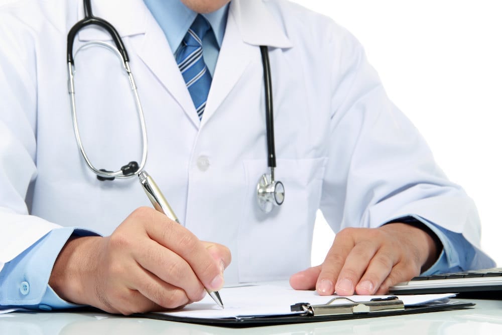 A doctor in a white coat writing on paper with a stethoscope around the neck